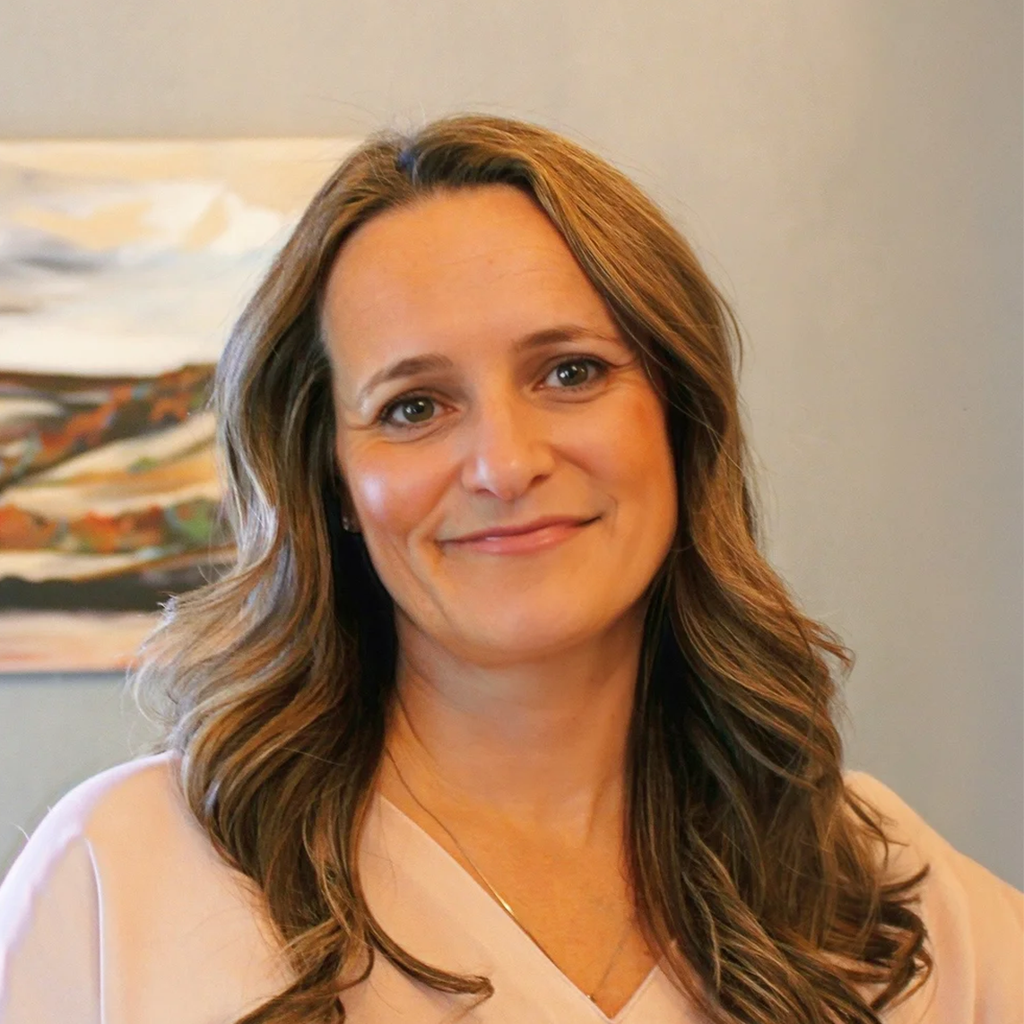 A woman with long, wavy brunette hair and light makeup smiling in an indoor setting, wearing a light pink blouse, with abstract artwork in the background.