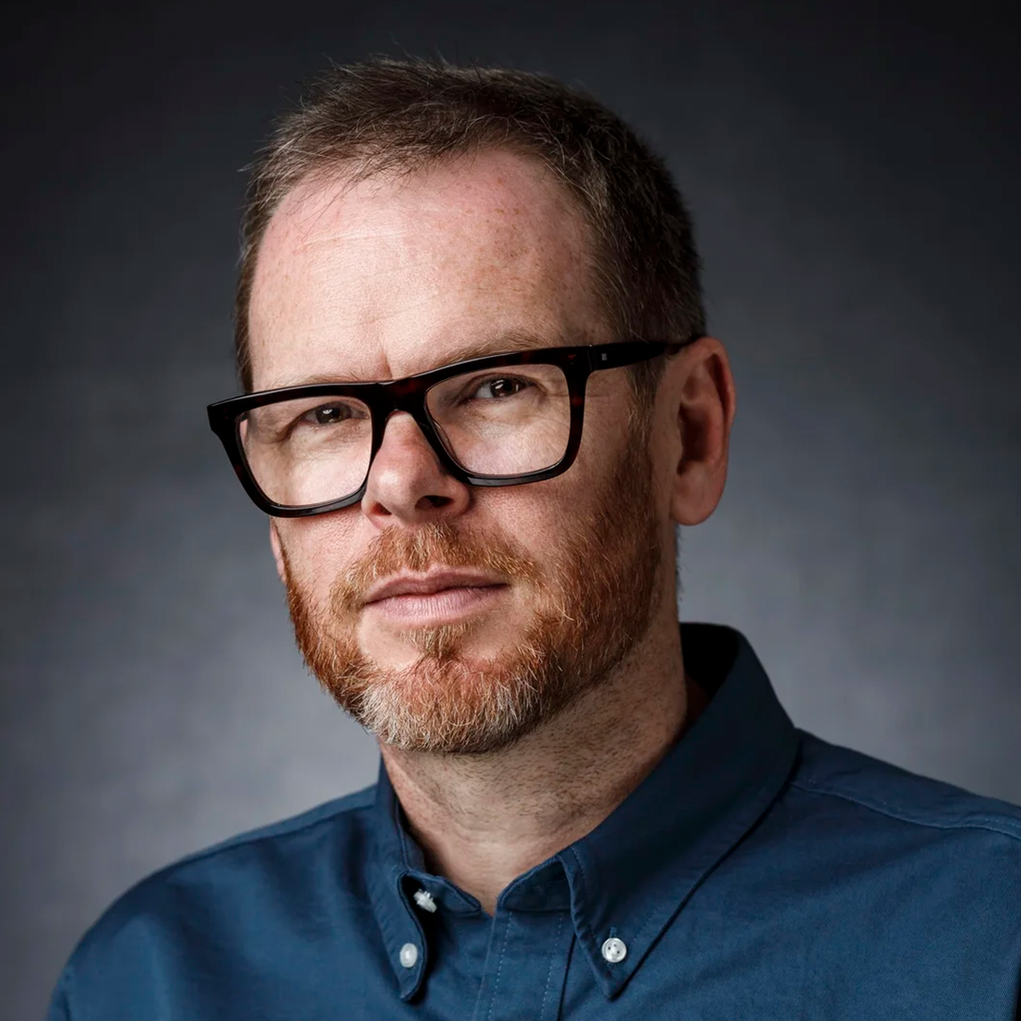 A man with short brown hair, glasses, and a beard, wearing a dark blue button-up shirt, looking at the camera against a dark gradient background.